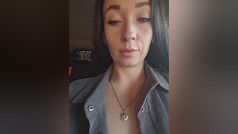 A video of a young woman with fair skin, long dark hair, and light makeup, wearing a button-down shirt. She has a serious expression. Background is dimly lit and blurred.