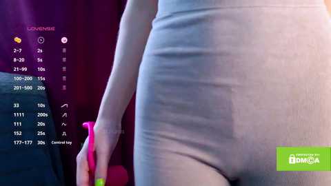 Video of a woman's torso in a tight, light-gray bodysuit. She holds a pink vibrator. Background features a dark curtain and digital clock display.