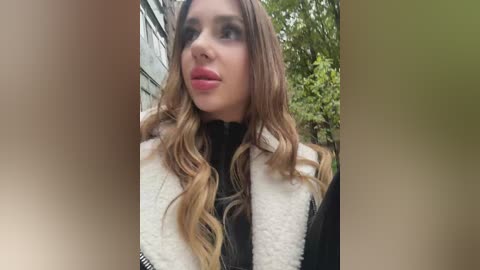 Media: A video of a young woman with long, wavy, light brown hair, wearing a black turtleneck and a white, textured coat, looking slightly to the left. Background shows blurred greenery and a building.