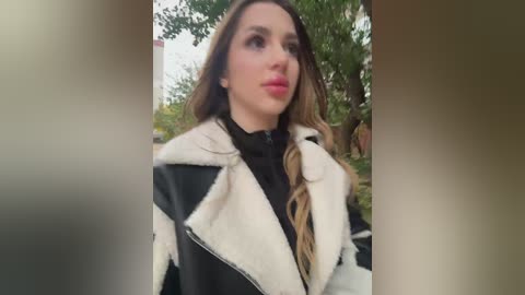 Media: Video of a young woman with long brown hair, wearing a black and white fur-trimmed coat, standing outdoors against a blurred greenery background.