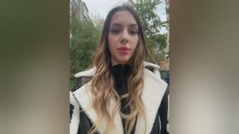 Media: Video of a young woman with long, wavy blonde hair, wearing a black turtleneck and white fur-trimmed jacket, standing outdoors with blurred greenery and urban buildings in the background.