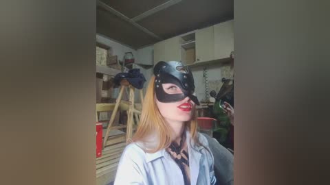 Video of a woman with long blonde hair wearing a black cat mask, red lipstick, and a light denim jacket, standing in a cluttered, dimly lit workshop with tools and boxes in the background.