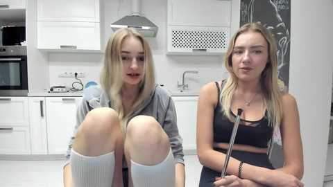 Media: Video of two young women in a modern kitchen. One has long blonde hair, grey hoodie, white knee-high socks, and bandaged knees. The other has blonde hair, black tank top, and holding a microphone.