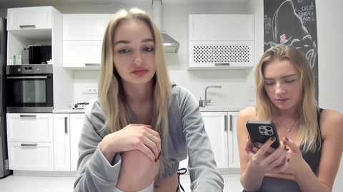 Media: Video of two young women in a modern kitchen: one in a gray sweatshirt, the other in a black tank top, both blonde, looking at a smartphone. White cabinets, stainless steel appliances, and a blackboard wall with chalk art in the background.