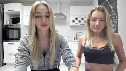 Media: Video of two young women, one with long blonde hair, wearing a gray hoodie, standing in a modern kitchen. The other has shorter hair, wearing a black crop top, standing beside her.