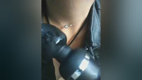 Close-up video of a person wearing a black leather jacket, holding a black camera, focusing on a necklace with a small pendant. The background is blurred, emphasizing the subject.