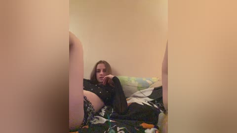 Video of a young woman with fair skin, long brown hair, and small breasts, lying on a messy bed with floral bedding, surrounded by beige walls.