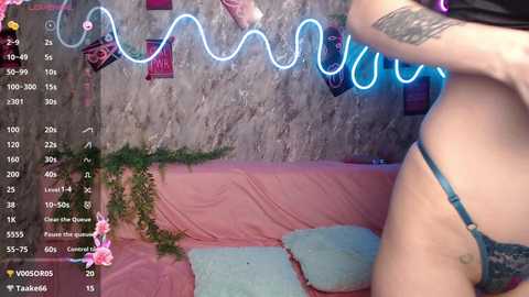 Video of a woman in blue lace lingerie, standing beside a pink couch, with a neon light background, showing her tattooed arm and thigh.