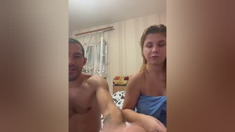 Video of a topless, smiling man with a light beard, and a woman with a towel wrapped around her shoulders, both sitting on a bed with beige walls and a window with white curtains.