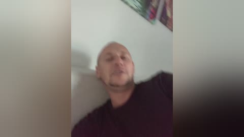 Media: A blurred video of a bald, middle-aged man with a beard, wearing a black shirt, leaning against a white wall with a colorful painting in the background.