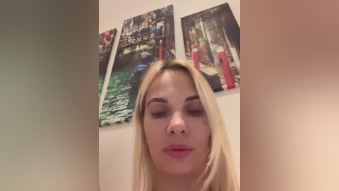 Media: A video of a blonde woman with closed eyes, wearing pink lipstick, standing in front of three colorful, nature-themed paintings on a beige wall.