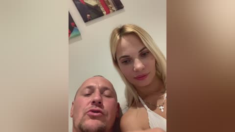 Media: Video of a smiling, blonde Caucasian woman with medium-sized breasts in a white dress, and a bald, Caucasian man with a trimmed beard, both indoors.