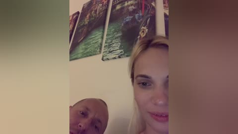 Media: A video captures a blonde woman with light skin and a man with a shaved head and dark skin, both smiling. Behind them, colorful paintings of boats on water hang on a beige wall.