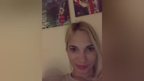 Media: A video of a blonde woman with light skin and a neutral expression, wearing a light-colored top, standing in a dimly lit room with a beige wall and two LEGO-themed posters in the background.