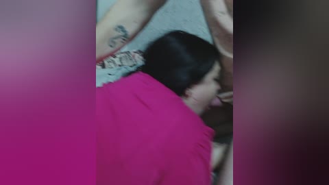 A blurred video showing a person with black hair and tattoos on their arms, lying on a pink surface, possibly a bed. The background is out of focus, with a neutral-colored wall. The image appears intimate and personal.