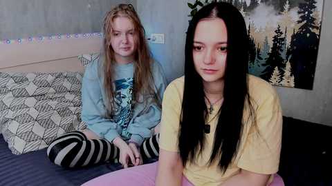 Video of two young women in a bedroom: one with long blonde hair in a blue hoodie, the other with long black hair in a yellow shirt.