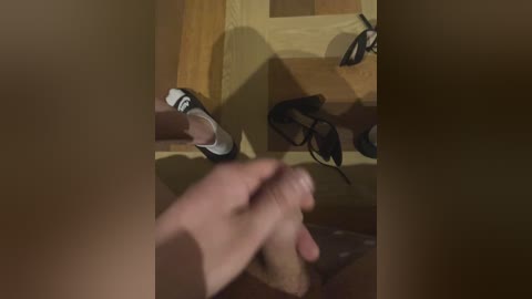 A video showing a hand holding a shoe, with a reflection of a person's legs in black high heels on a wooden floor, and a blurred person sitting on a couch in the background.