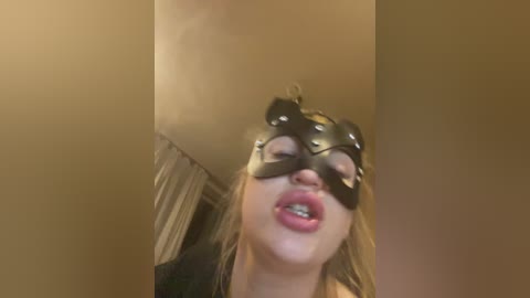 Video of a blonde woman with a black mask, looking seductively with parted lips, in a dimly lit room with beige walls and curtains.