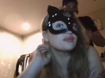 Video of a woman with long, blonde hair, wearing a black cat mask, sitting on a bed in a dimly lit room.