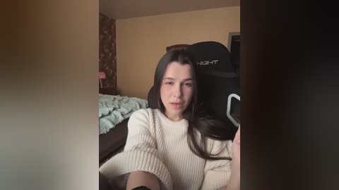 Video of a young woman with long dark hair, wearing a beige sweater, seated on a black gaming chair in a dimly lit room with a messy bed and floral-patterned wall.