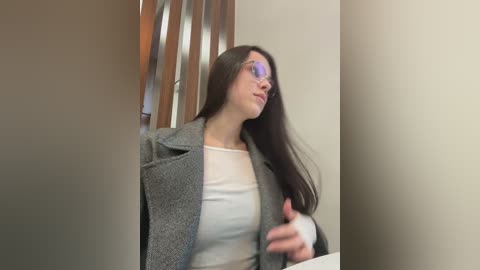 Video of a young woman with long dark hair, wearing a gray coat over a white top, seated in a wooden chair, gesturing with her right hand.