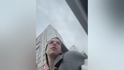 Video of a young woman with glasses, wearing a gray blazer, gazing upwards at a tall, modern skyscraper under a cloudy sky.
