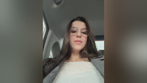A video of a young woman with long brown hair, wearing glasses, a white top, and gray cardigan, seated in a car, with blurred background.