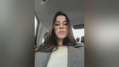Video of a young woman with long brown hair, wearing glasses, a gray blazer over a white shirt, sitting in a car with neutral interior.