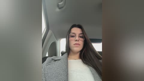 Video of a young woman with long dark hair, wearing glasses, gray jacket, and white top, sitting in a car, with a blurred background.