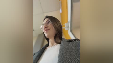 A video of a young woman with glasses, light skin, and straight brown hair, wearing a white shirt and gray cardigan, standing in a beige and yellow tiled bathroom.