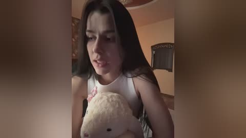 A video of a young woman with long black hair, wearing a white tank top, holding a plush teddy bear, indoors with warm lighting and blurred background.