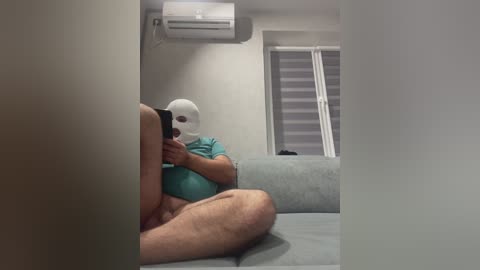 Video of a person wearing a white mask and turquoise shirt, sitting on a light blue sofa, holding a phone, with an air conditioner above and blinds in the background.