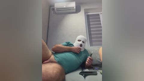Video of a man in a white mask, green shirt, and no pants, lying on a gray couch, holding a phone. Background includes an air conditioner, blinds, and a beige pillow.