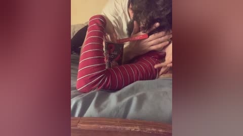 Video of a woman with curly hair wearing red and white striped pajamas, lying on a bed with blue sheets. She is sucking on a red lollipop, with her hand on her chest, and a man's arm partially visible.