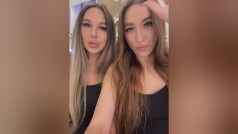 Media: Video of two young women with long, straight blonde hair, fair skin, and wearing black tank tops. They are standing close together, smiling, and looking directly at the camera.