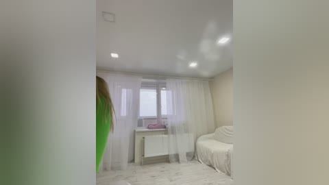 Video of a minimalist, bright bedroom with white walls, a beige sofa, white curtains, a radiator, and a green scarf in the foreground.