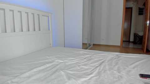 Media: Video of a minimalist, modern bedroom with a white wooden headboard, neatly made white bed, and a large mirror reflecting a doorway and wooden floor.