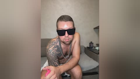 A video of a shirtless, muscular man with tattoos, wearing black sunglasses and a silver chain necklace, seated on a bed in a dimly lit room.