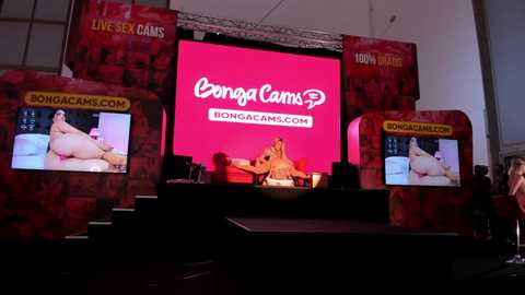 Media: Video of a live sex cam event featuring a voluptuous woman with fair skin and blonde hair, reclining on a bed, displayed on a large red backdrop.