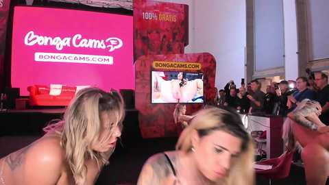 Media: Video of two women in lingerie, one blonde with wet hair, the other with blonde hair, standing in front of a red CamSoda banner and a large pornographic screen in a dimly lit room filled with seated men.