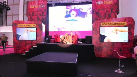 Media: Video of a vibrant, red-themed Bongacam booth at a convention, featuring two large screens with erotic content, a podium, and a stool.