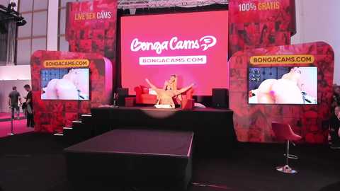 Media: Video of a vibrant, red-themed booth at a convention, featuring two large screens showcasing adult content, a podium with a model, and a banner reading \"BongaCams.com.\