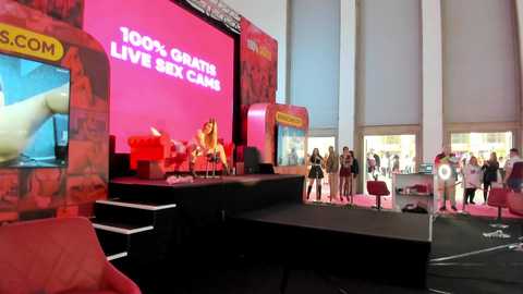 Media: Video of a vibrant stage set with a large pink screen displaying \"100% LIVE SEX CAMS\" in white letters. Audience members stand in front, some holding signs.