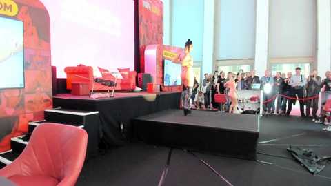 Media: Video of a vibrant, modern conference room with a large screen, red furniture, and a presenter in a yellow outfit on a stage.