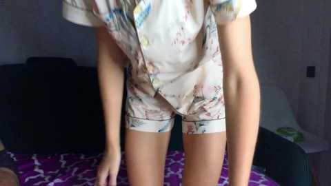 A video of a person wearing floral pajamas, leaning forward on a purple-patterned bed. The background includes a black couch and a white lamp.