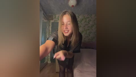 Video of a smiling blonde woman in a black top and black tights reaching out in a dimly lit, wallpapered bedroom with a bed and wardrobe.