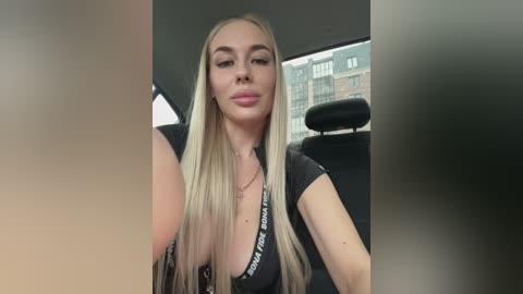 Video of a blonde woman with long hair, fair skin, and a slender physique, wearing a black top inside a car, with blurred buildings visible through the window.