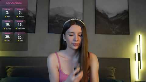 Media: Video of a young woman with long, straight brown hair, wearing a pink camisole, sitting on a bed in a dimly lit room with black-and-white mountain scenery artwork.