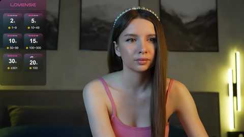 Media: Video of a young woman with long brown hair, wearing a pink tank top, seated indoors. Background features a gray headboard, two framed black-and-white photos, and a glowing lamp.