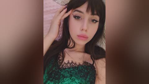 A video of a young woman with long black hair, fair skin, and brown eyes, wearing a green lace camisole. She lies on a soft, light-colored bedspread, her hand gently touching her hair.